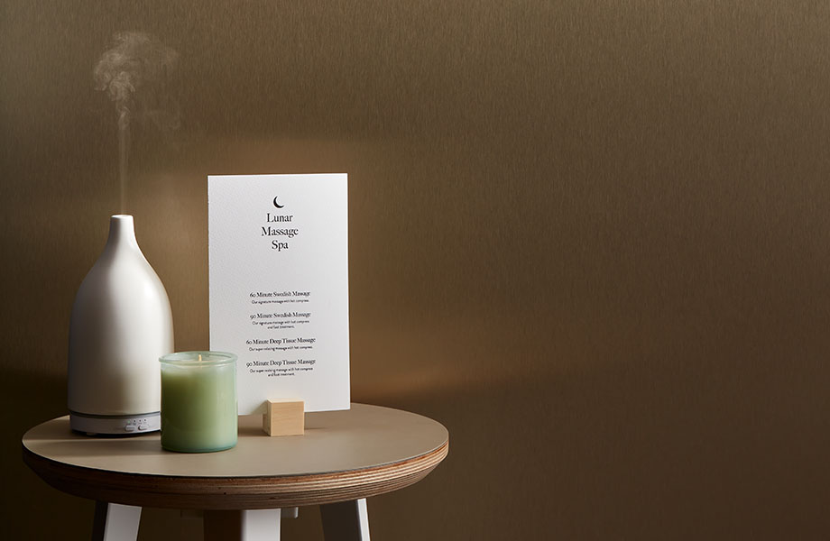 Round side table in a spa with candle, diffuser and list of services. Back wall is made of bronze metal laminate.
