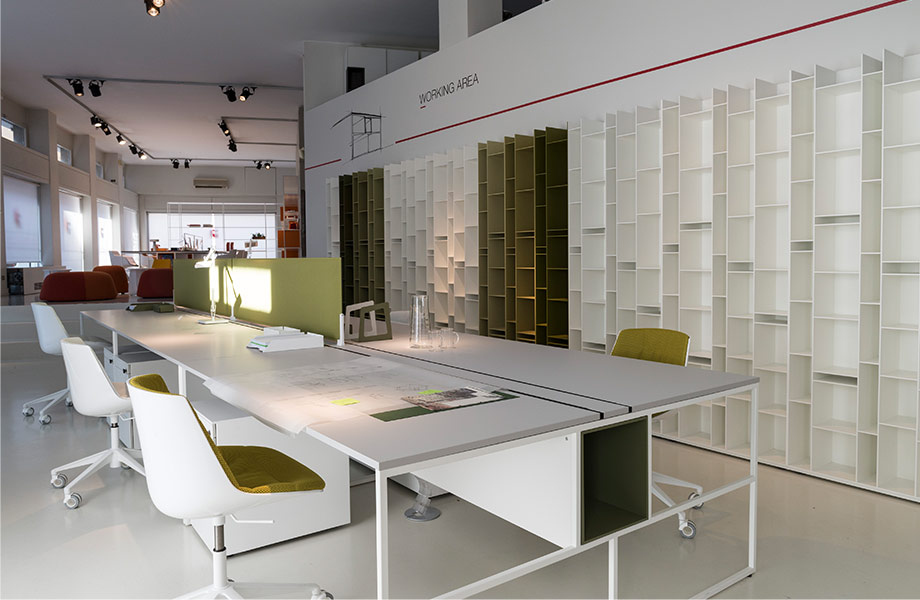 Open communal office workspace with white tabletop, white and green chairs, and wall of shelving