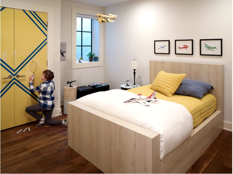 Chambre d’enfant avec un enfant qui écrit sur les portes de la garde-robe en stratifié de marque Formica Jaune Chromé et Bleu Matrice avec une base de lit double en stratifié de marque Formica Planche de Chêne Brut avec literie jaune, bleue et blanche.